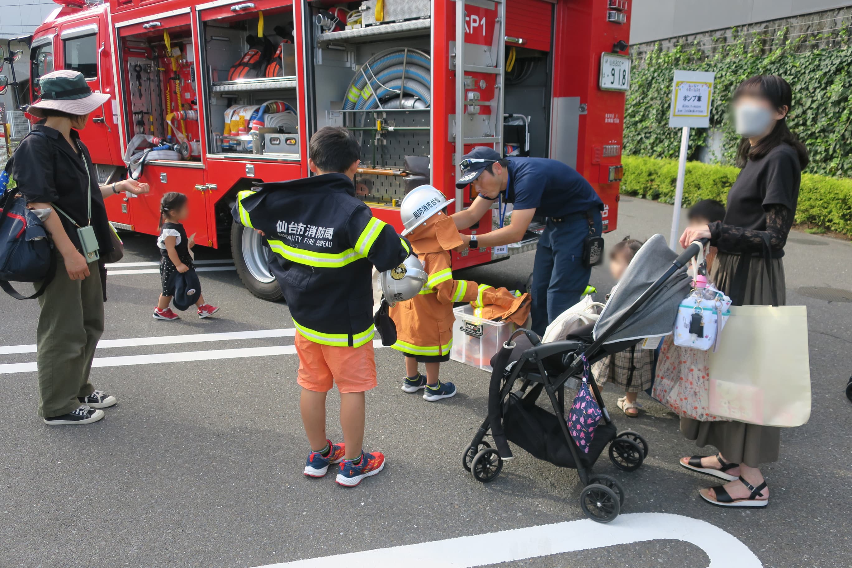 展示されている消防車両の脇で、消防団員から消防服を着させられる男の子